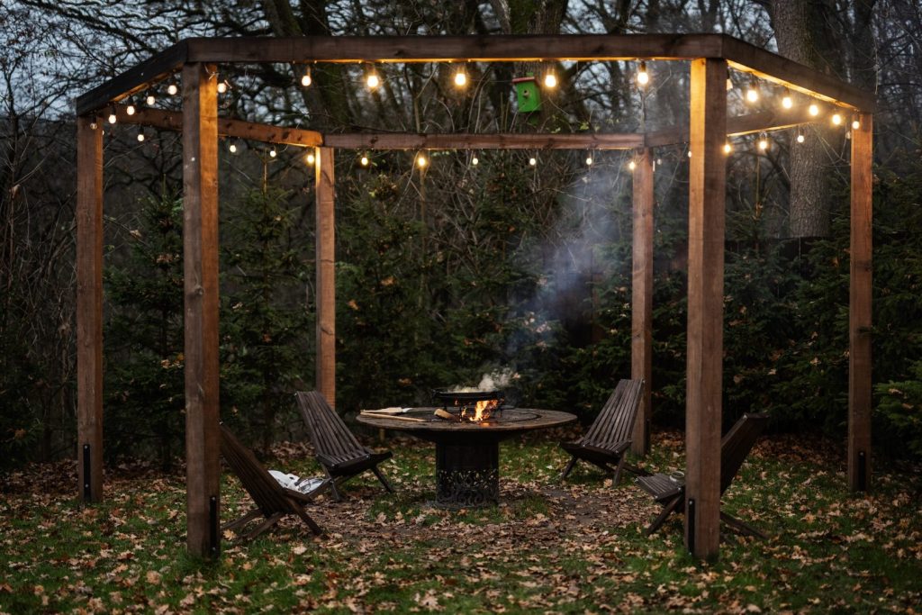 Wooden garden pergola with grill at twilight, decor lamp and chairs in autumn.